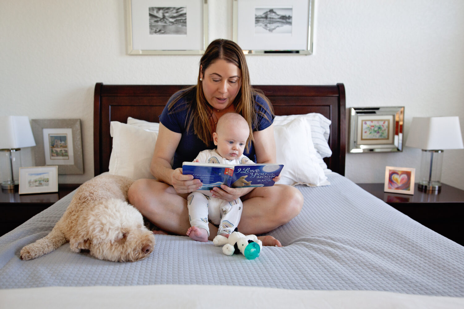 Mom reading to her baby boy on the bed with the dog napping next to them.